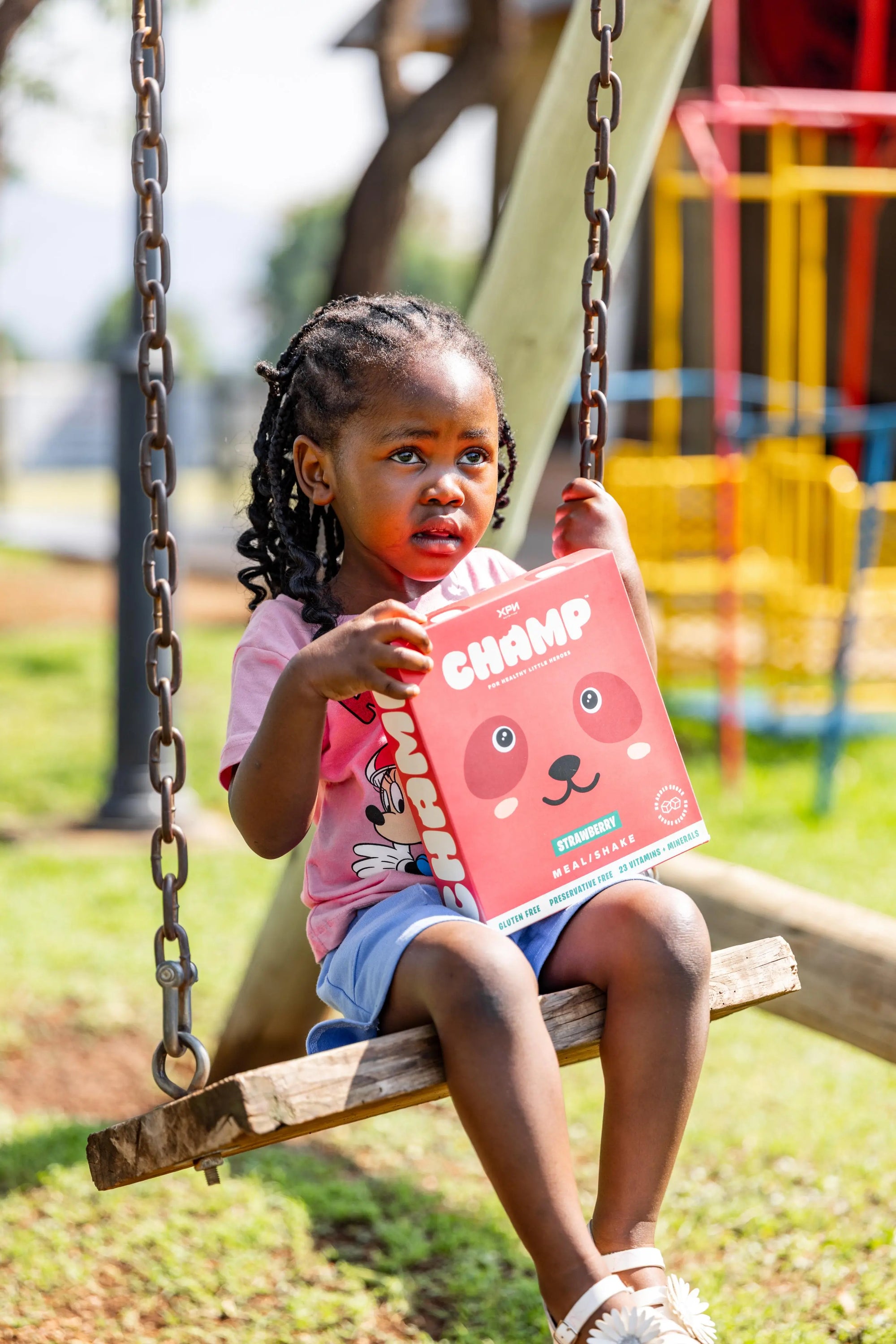 Child on a swing holding a 'Champ' nutrition box in an outdoor playground setting.